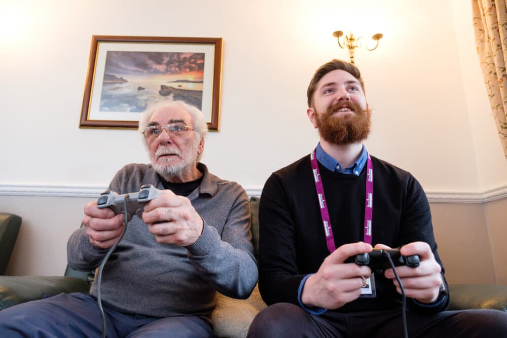 Two men, an elderly client and a Home Instead Care Professional, sitting on a couch playing video games and smiling. - Home Instead Bournemouth & Christchurch