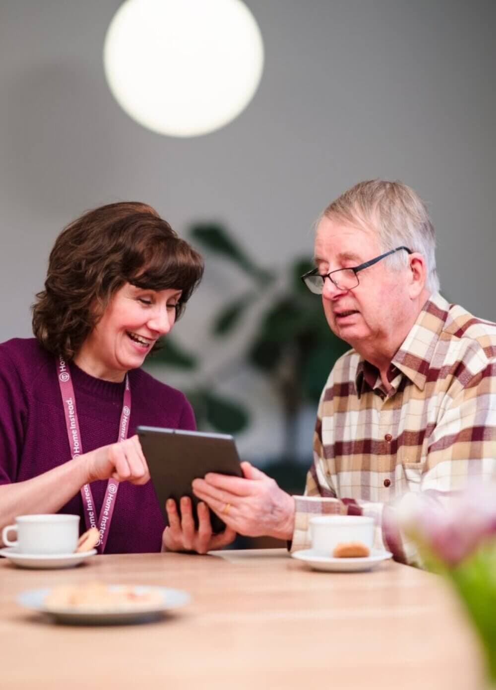Two women sitting on a couch, smiling and holding hands, with tea and a tray on the table in front of them. - Home Instead Bournemouth & Christchurch
