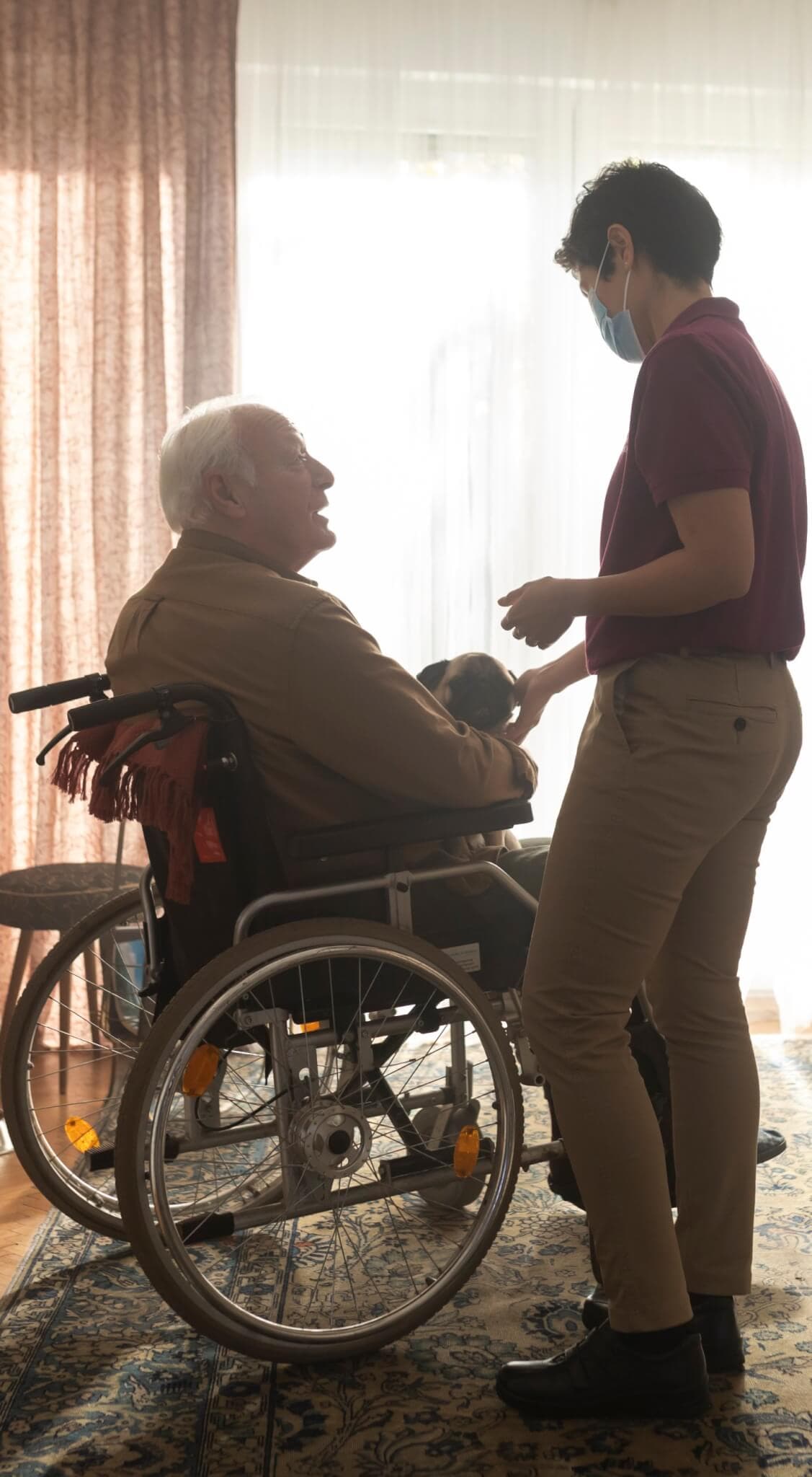 A person in a wheelchair holding a small dog is attended to by a standing caregiver wearing a face mask. - Home Instead