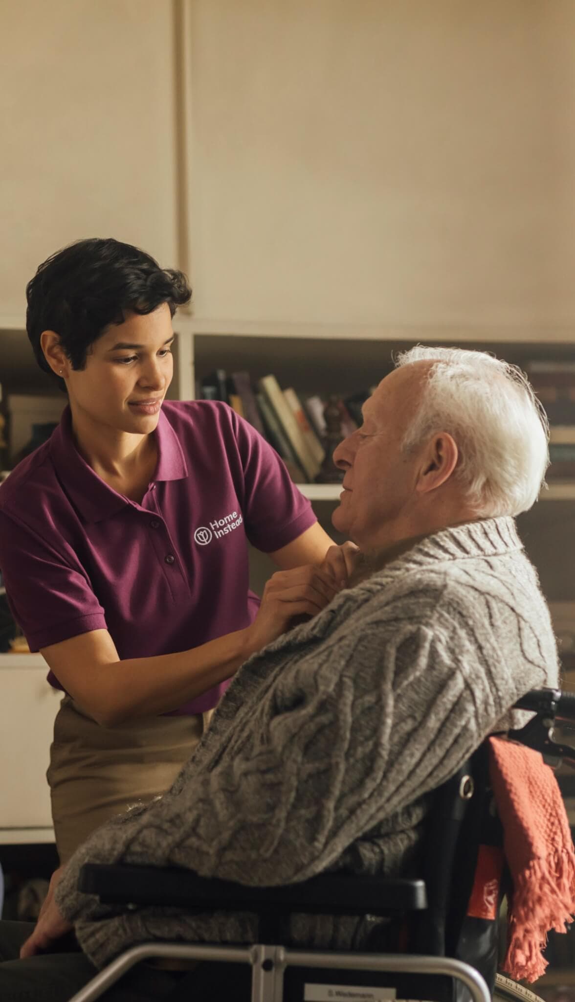 A caregiver in a purple shirt helps an elderly man in a wheelchair with his sweater in a cozy room. - Home Instead