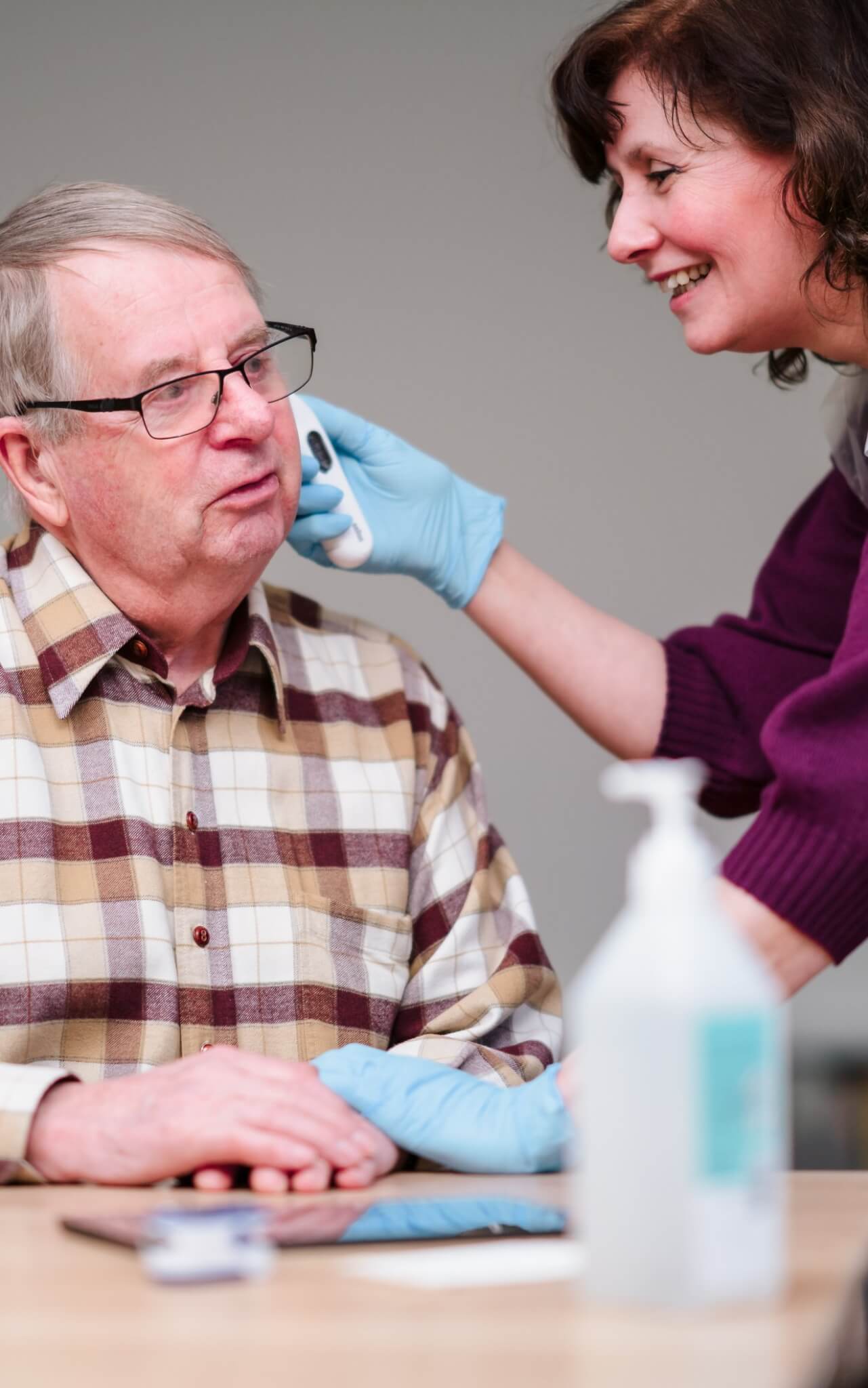 A woman wearing gloves helps an older man with glasses by holding a cloth or device to his face. - Home Instead