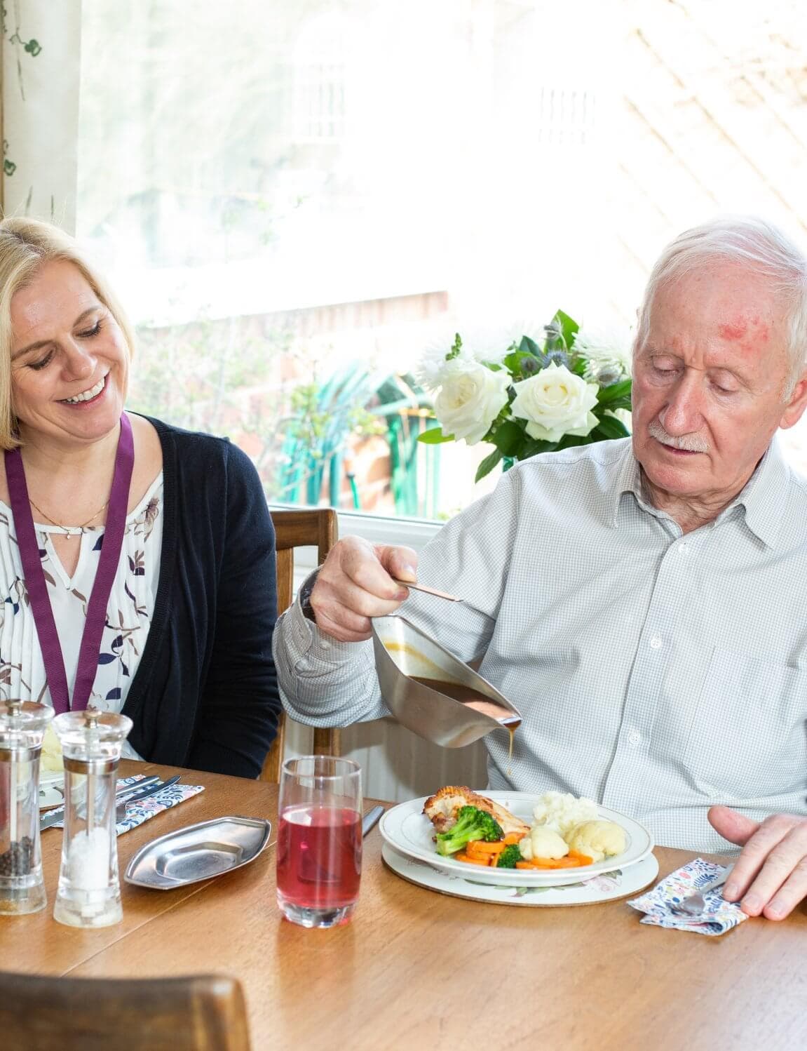 A smiling woman sits next to an older man pouring gravy onto his meal at a dining table. - Home Instead