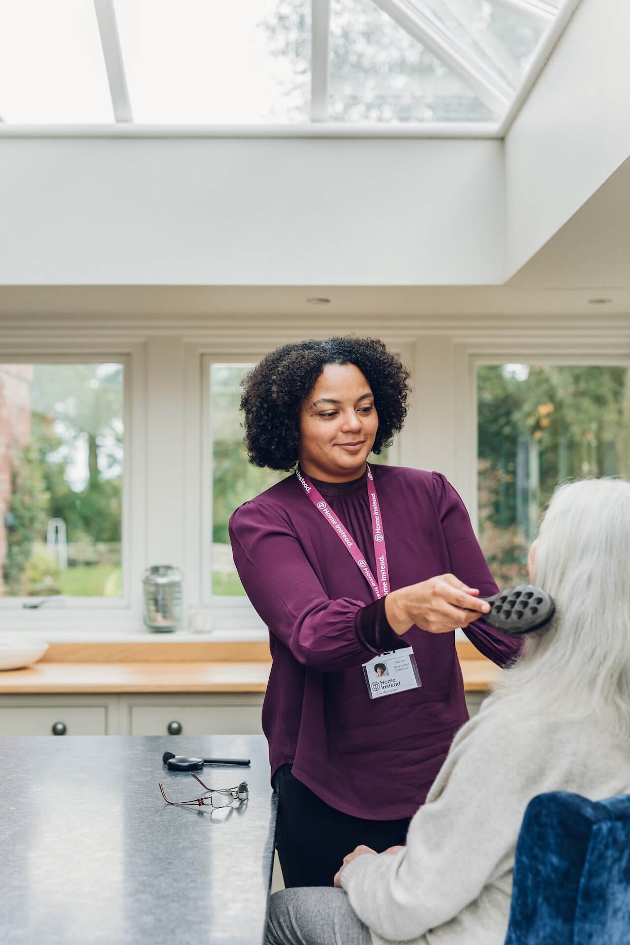 A caregiver with a name badge brushes the hair of an elderly woman sitting in a bright, modern kitchen. - Home Instead