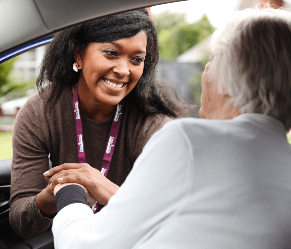A smiling caregiver with a lanyard helps an elderly person into a car. - Home Instead