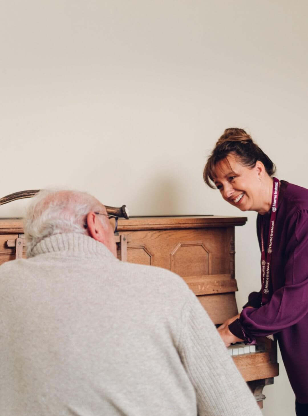 A woman in a purple shirt smiles while playing the piano for an elderly man in a gray sweater. - Home Instead