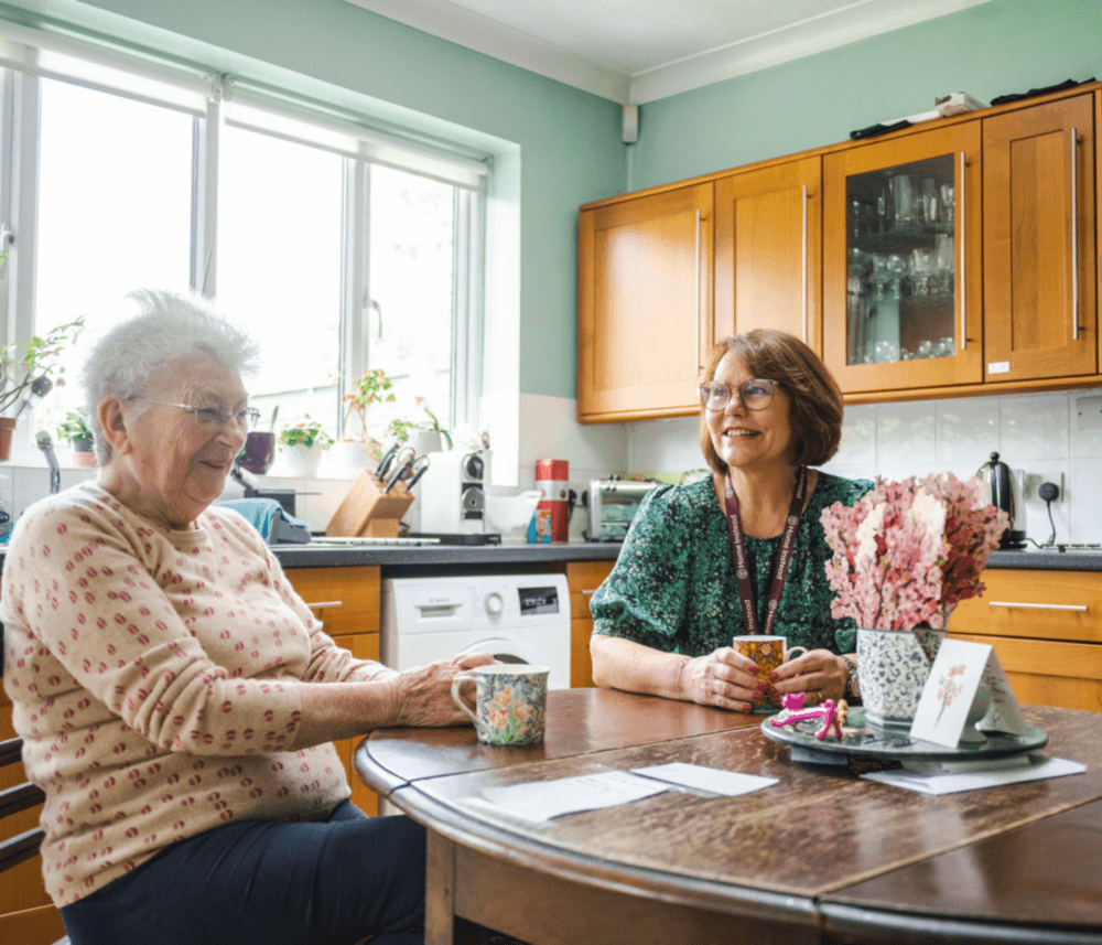 Two women sitting at a kitchen table, smiling and holding mugs, with flowers and cards on the table. - Home Instead