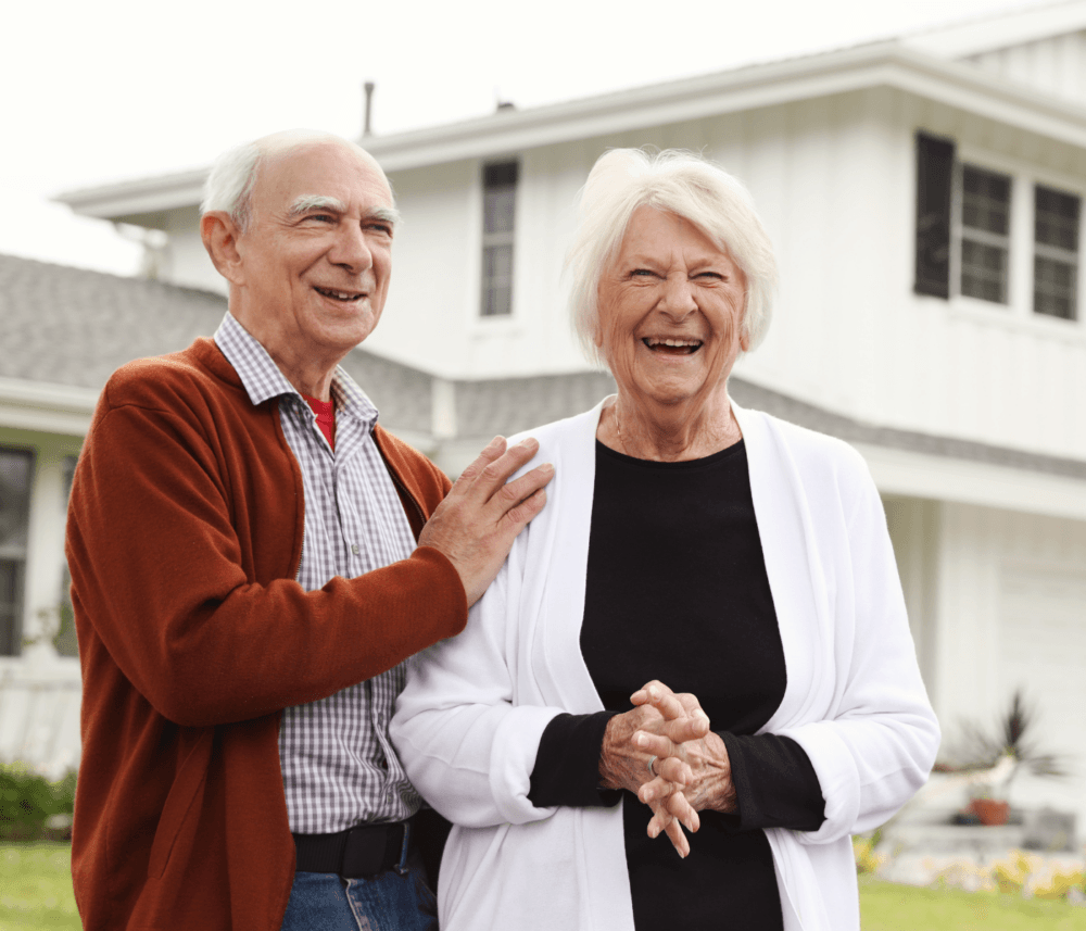 Older couple smiling and standing together in front of a white house on a sunny day. - Home Instead