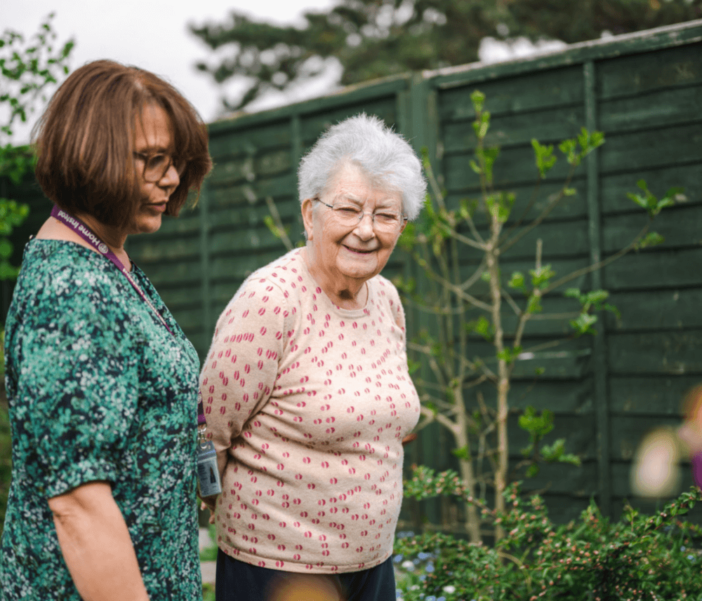 Two women, one elderly and one middle-aged, standing in a garden with a green fence and plants in the background. - Home Instead