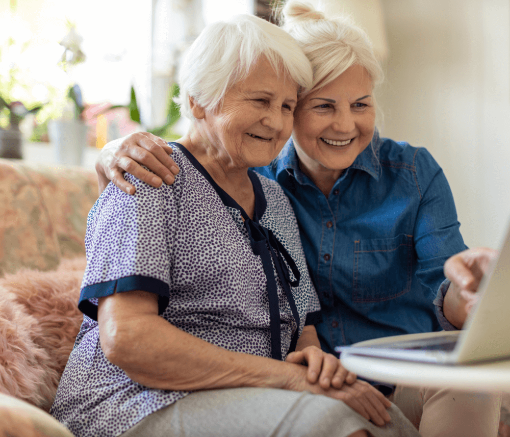 Two elderly women sitting together on a couch, smiling and looking at a laptop screen. - Home Instead