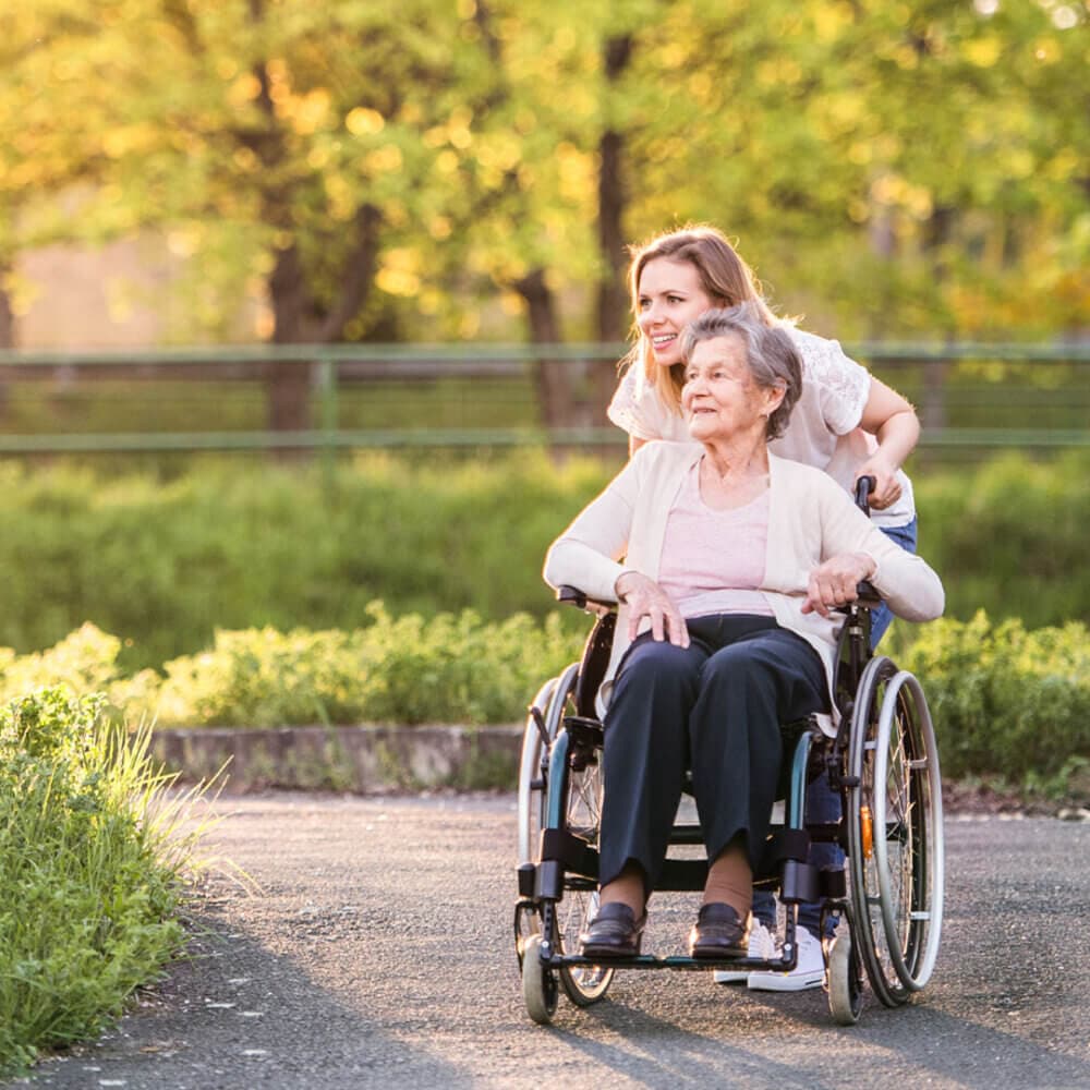A young woman smiles while pushing an older woman in a wheelchair along a sunny park path. - Home Instead