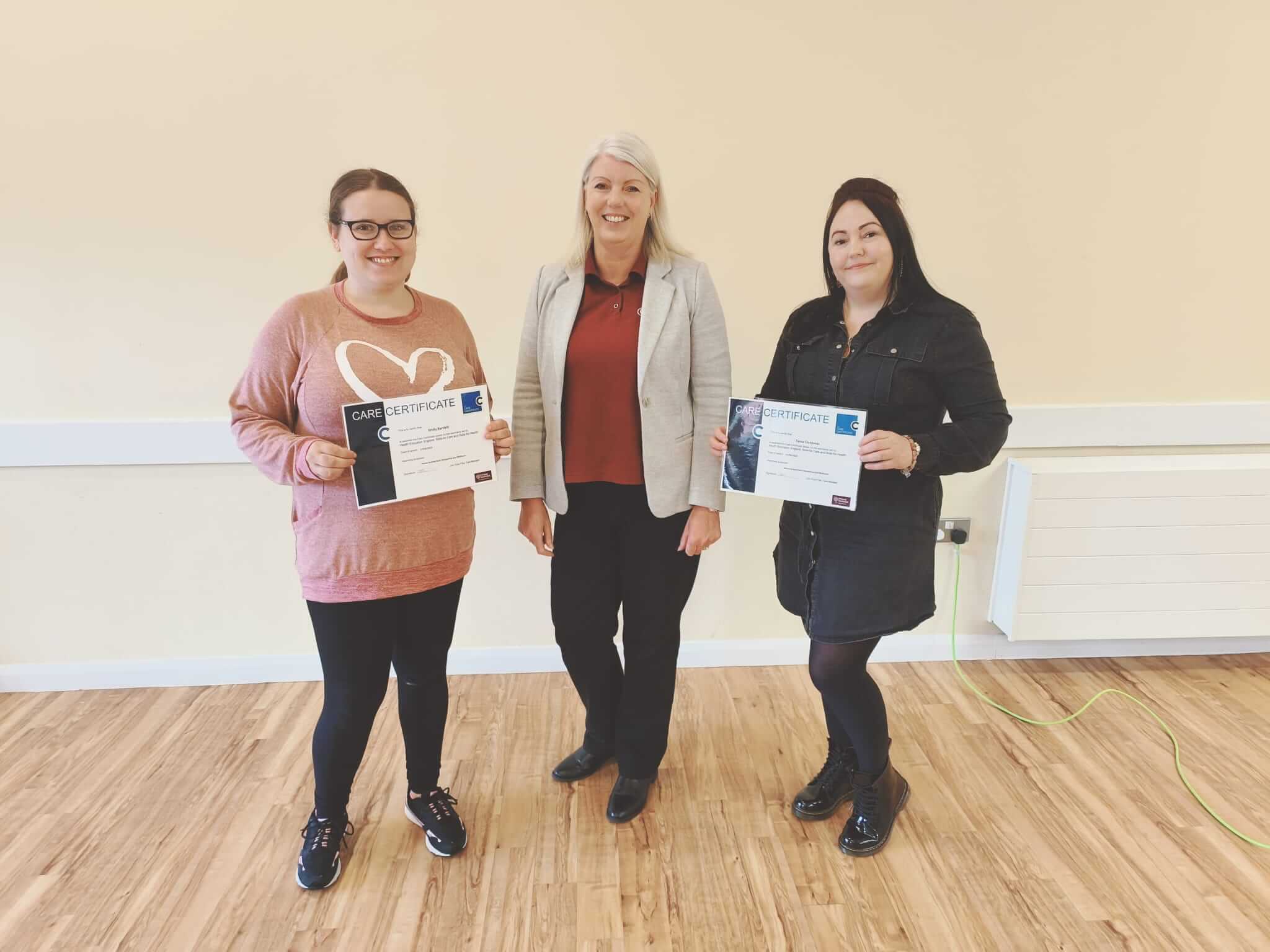 Three women are standing. The two outer women hold certificates while the woman in the center stands without one, smiling. - Home Instead
