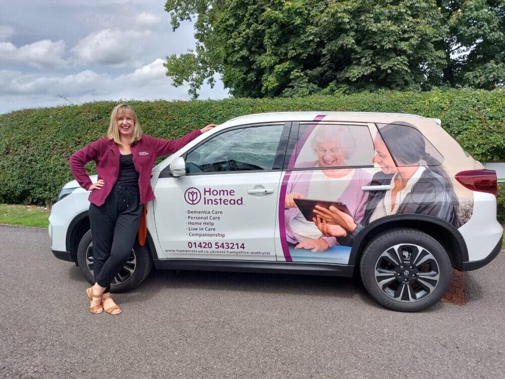 A woman in a maroon jacket leans on a Home Instead Healthcare car with service details on the side. - Home Instead