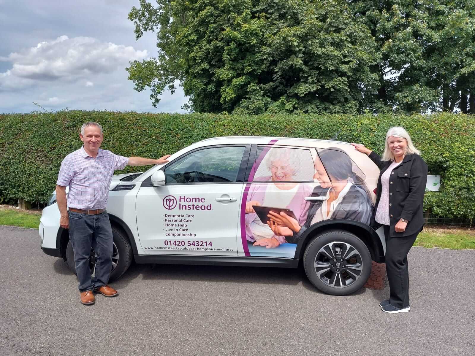 Two people stand beside a car with "Home Instead" branding, featuring images related to elder care services. - Home Instead