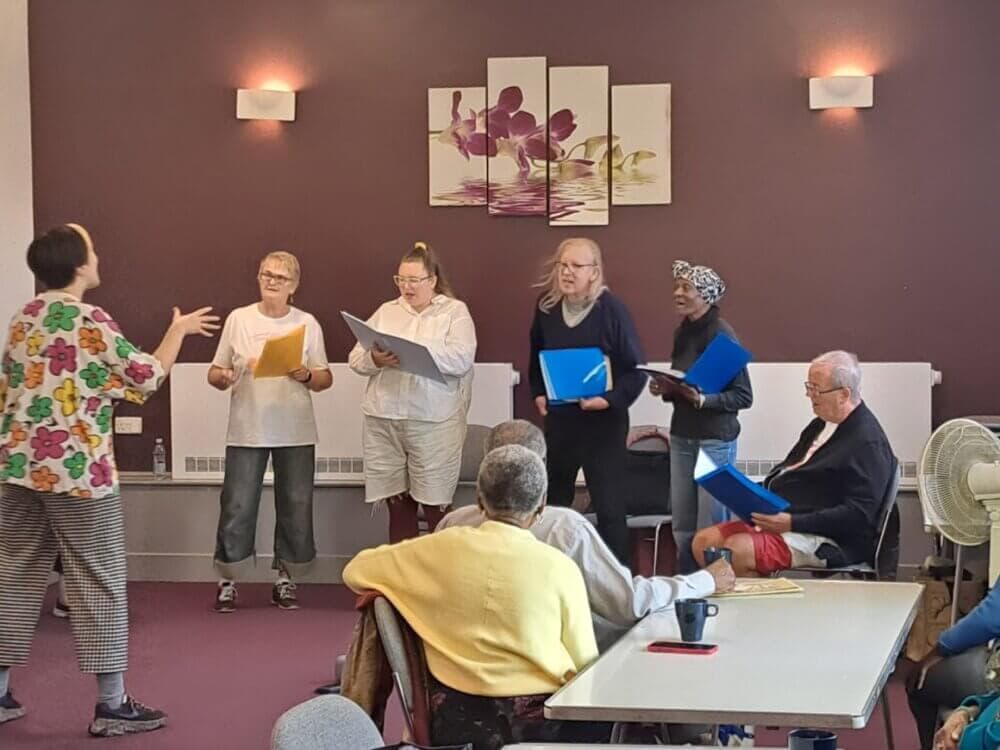 A group of people standing and holding folders, engaging in an activity, while another person gestures toward them. - Home Instead