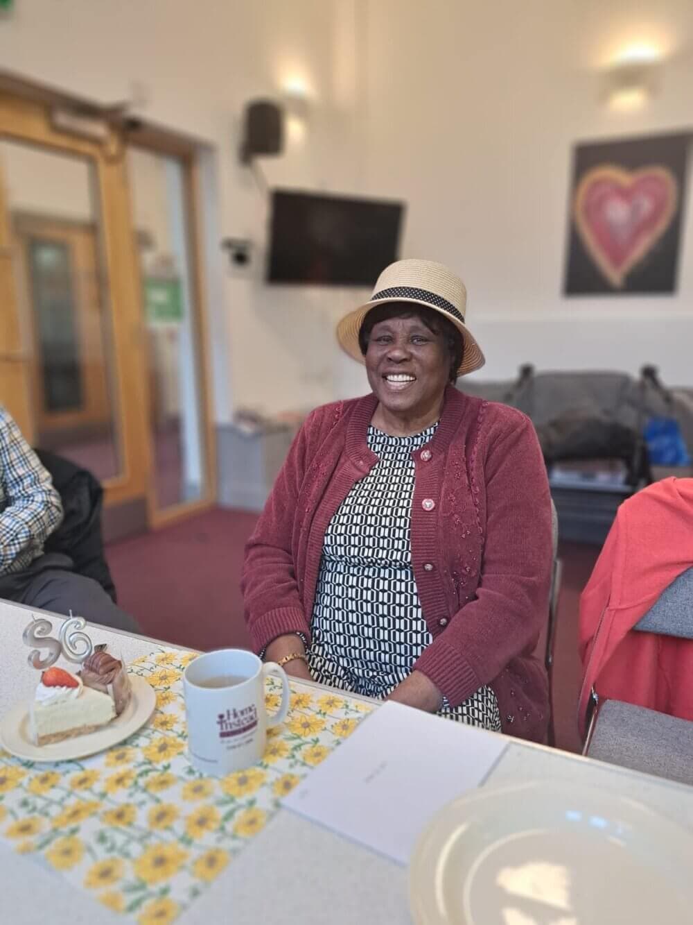 A smiling woman wearing a hat, sitting at a table with a cake and birthday candles showing the number 66. - Home Instead