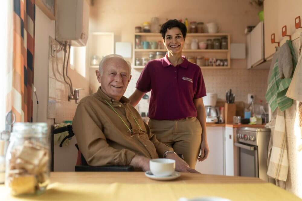 An elderly man in a wheelchair and a woman standing beside him smile in a cozy kitchen setting. - Home Instead