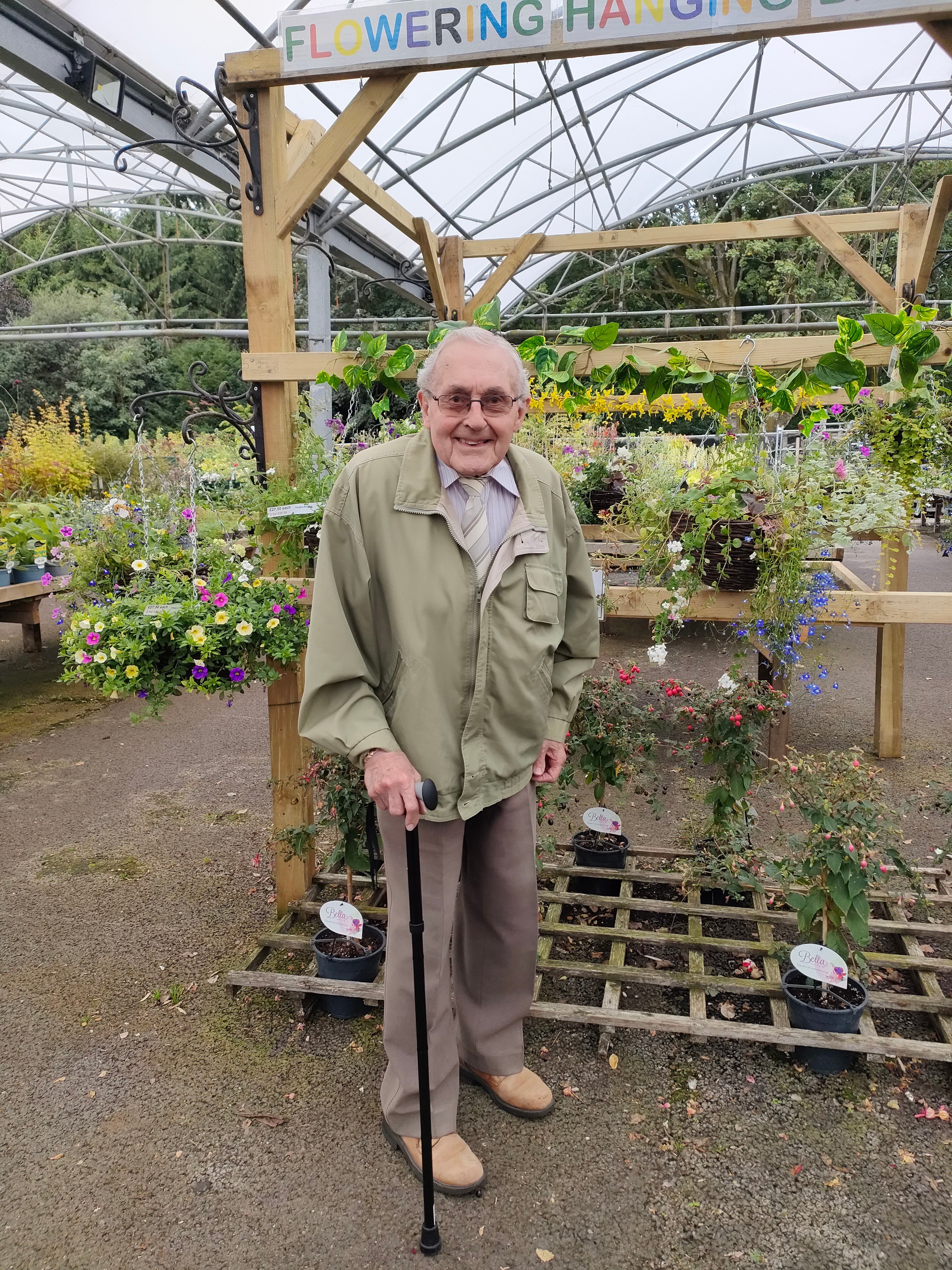An elderly man with a cane stands in a garden center surrounded by flowering plants and hanging baskets. - Home Instead