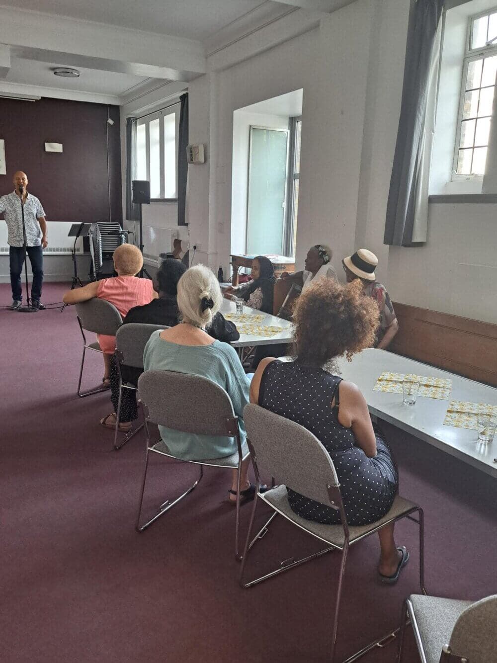 A man is speaking to a group of seated people in a room with large windows and a maroon carpet. - Home Instead