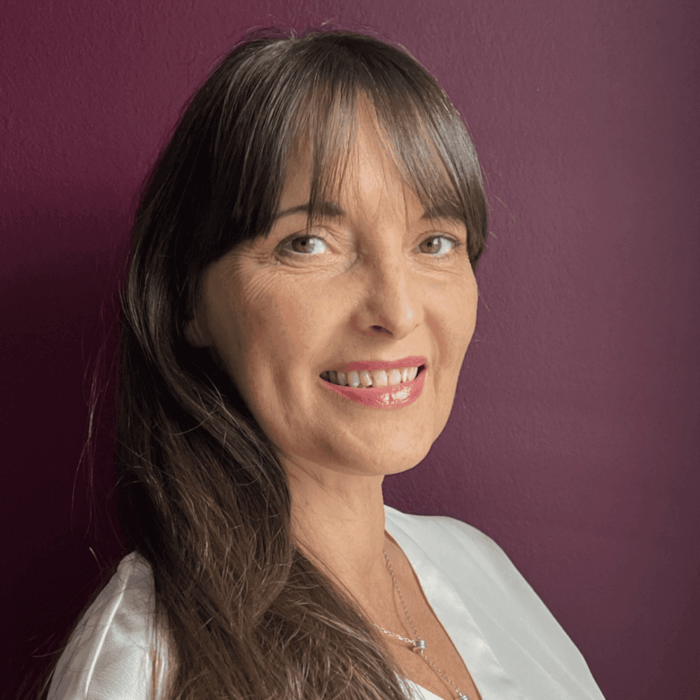 A woman with long brown hair and bangs, smiling, wearing a white top, in front of a burgundy background. - Home Instead