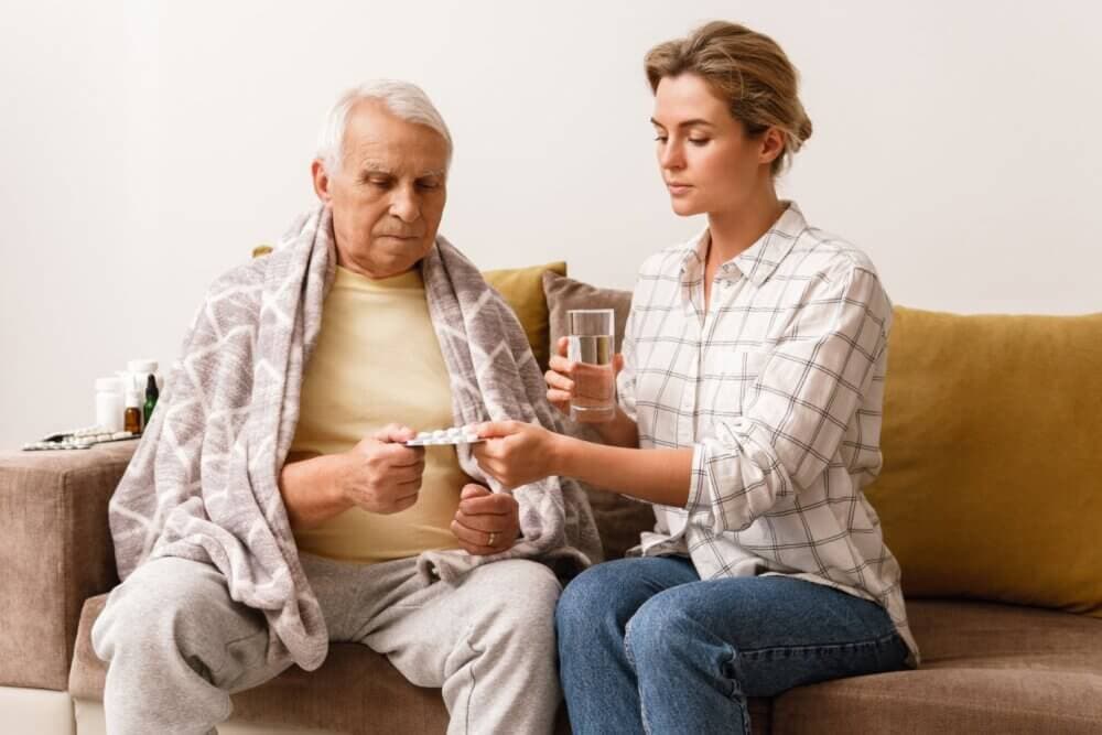 Young woman helps elderly man with medication, offering water and pills on a cozy couch. - Home Instead