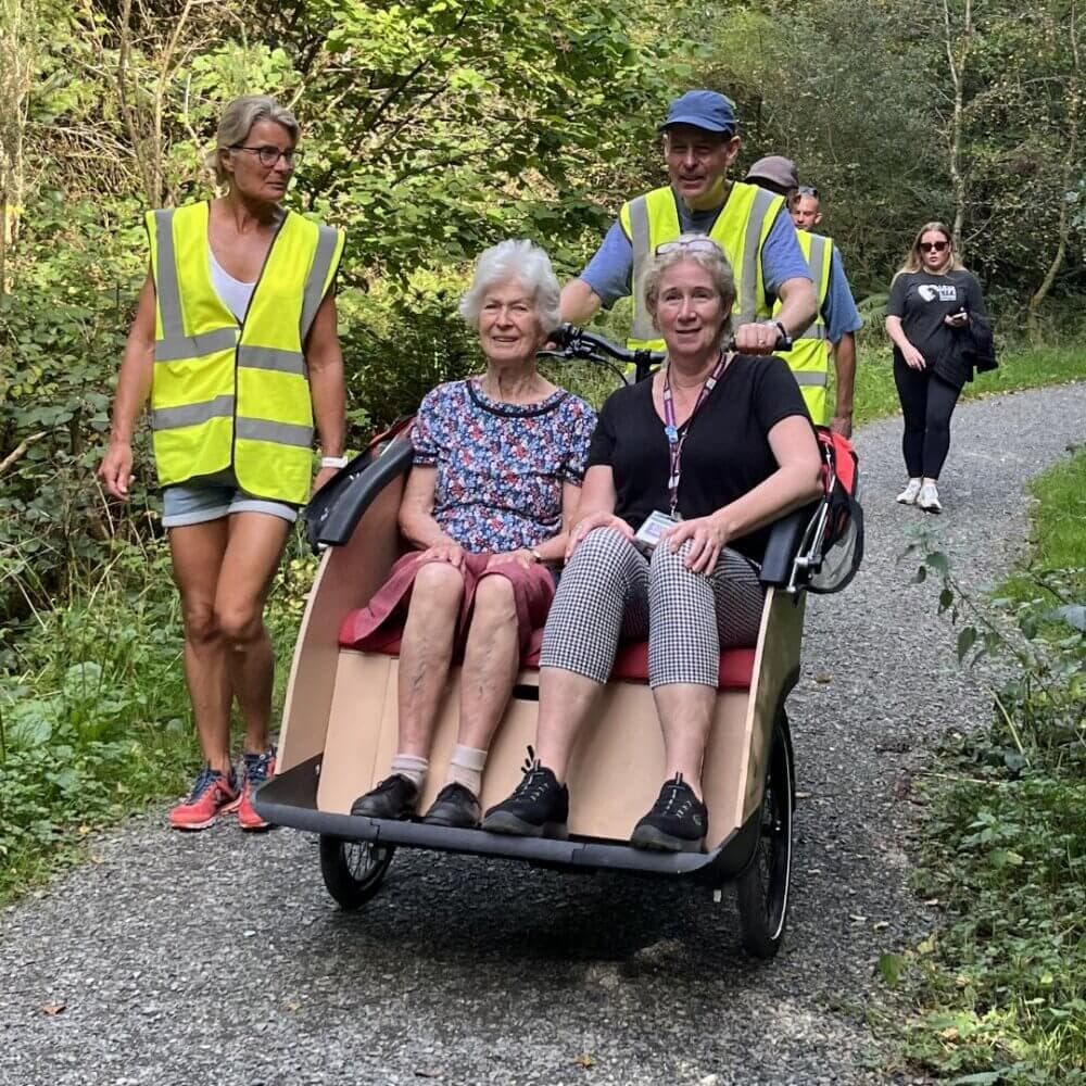 Two women are seated in a rickshaw, being pushed by a man; another woman walks alongside on a forest trail. - Home Instead