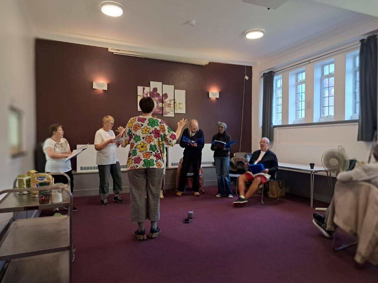A group of people standing and singing in a room, with sheet music, directed by a person in a floral shirt. - Home Instead
