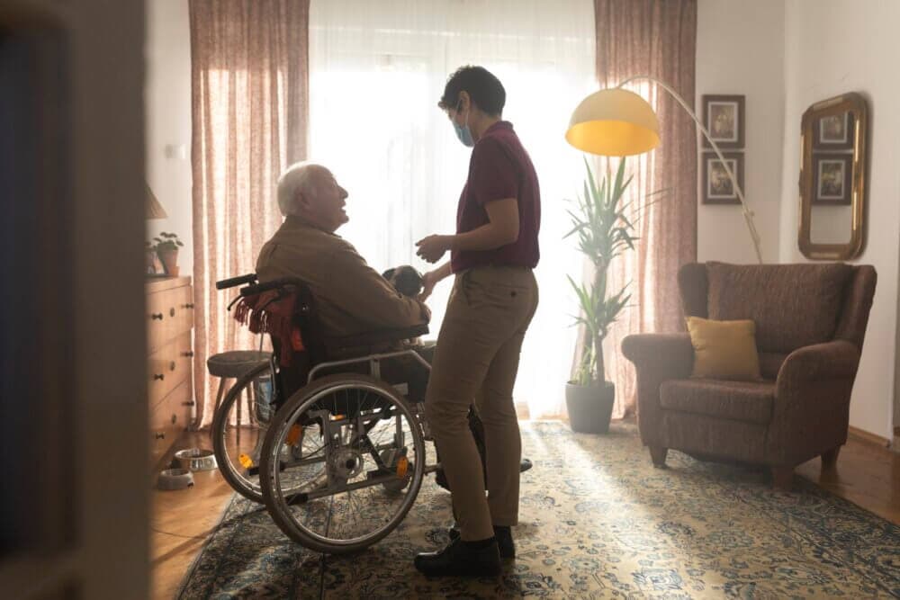 A caregiver speaks to an elderly man in a wheelchair in a cozy living room with natural light and warm decor. - Home Instead