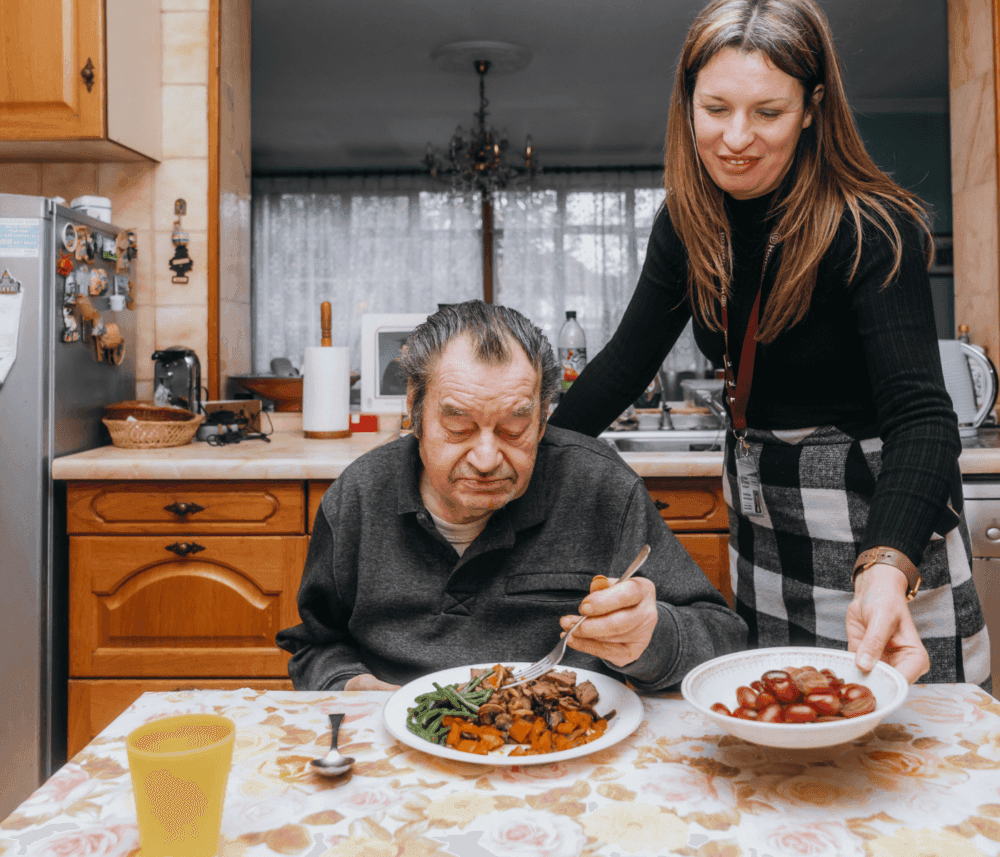A caregiver serves grapes to an elderly man at a kitchen table with a plate of food in front of him. - Home Instead