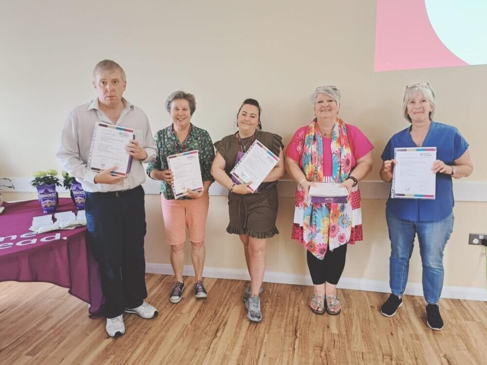 Five people standing indoors and holding certificates, smiling at the camera. A table with items is visible on the left. - Home Instead