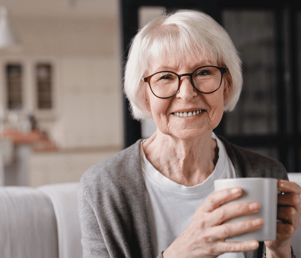 Smiling elderly woman with short white hair and glasses holding a white mug, wearing a light sweater indoors. - Home Instead