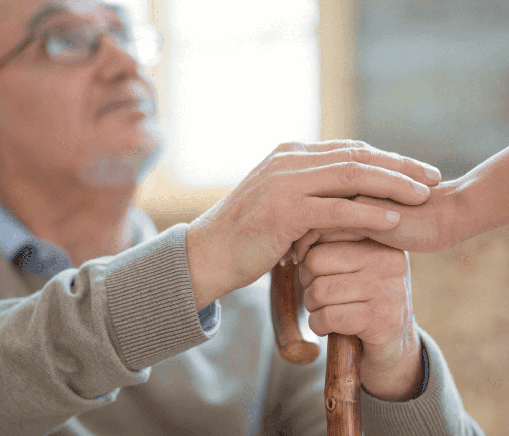 Elderly man holding a wooden cane, another person’s hand offering support. - Home Instead