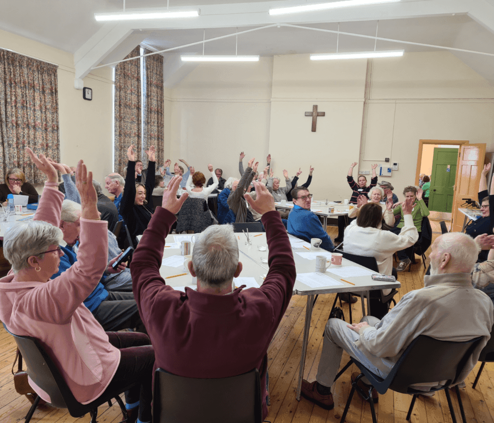 A group of elderly people in a bright room with a cross, seated in a circle with hands raised, engaging in a cheerful activity. - Home Instead