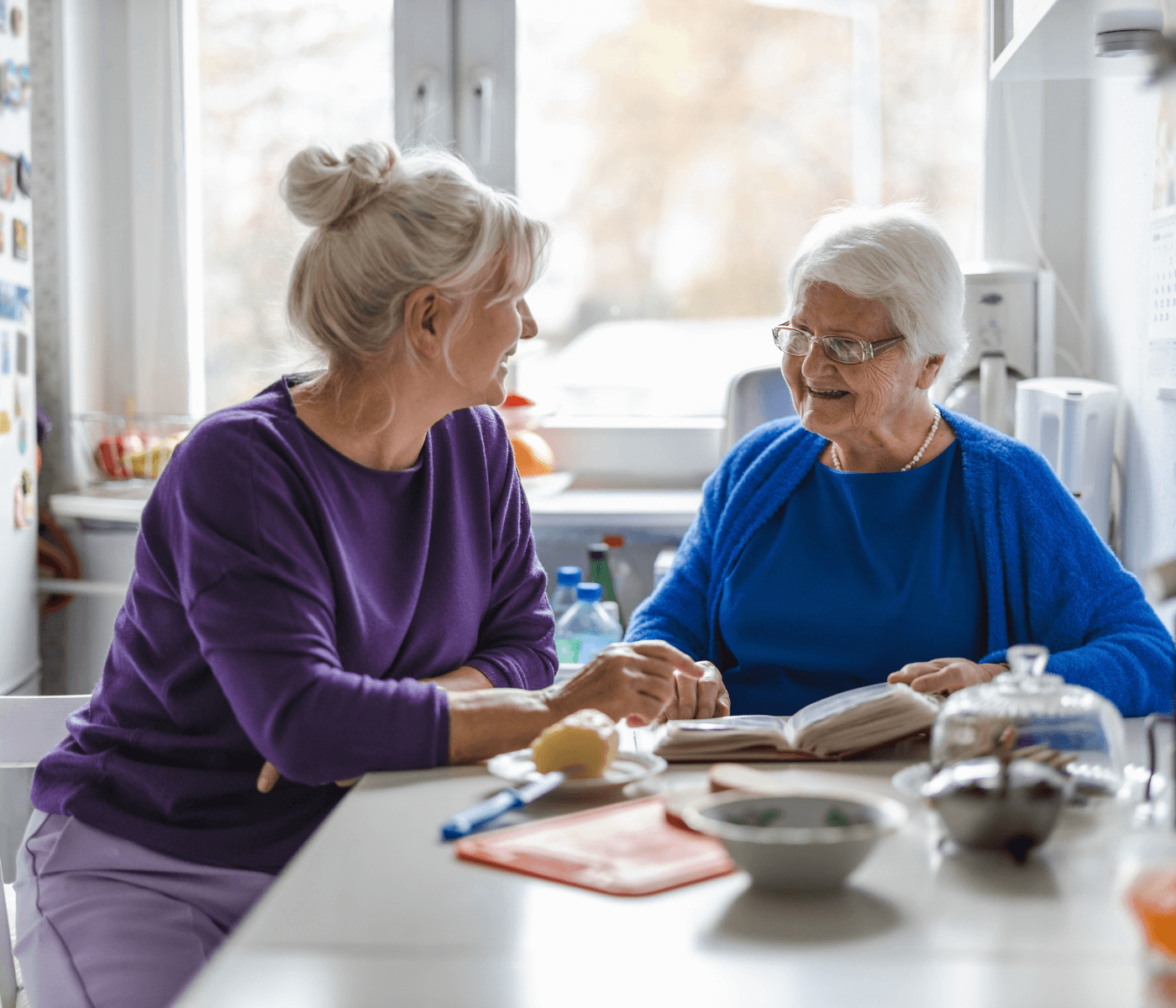 Two elderly women with grey hair smiling and talking at a kitchen table, one holding a book. - Home Instead