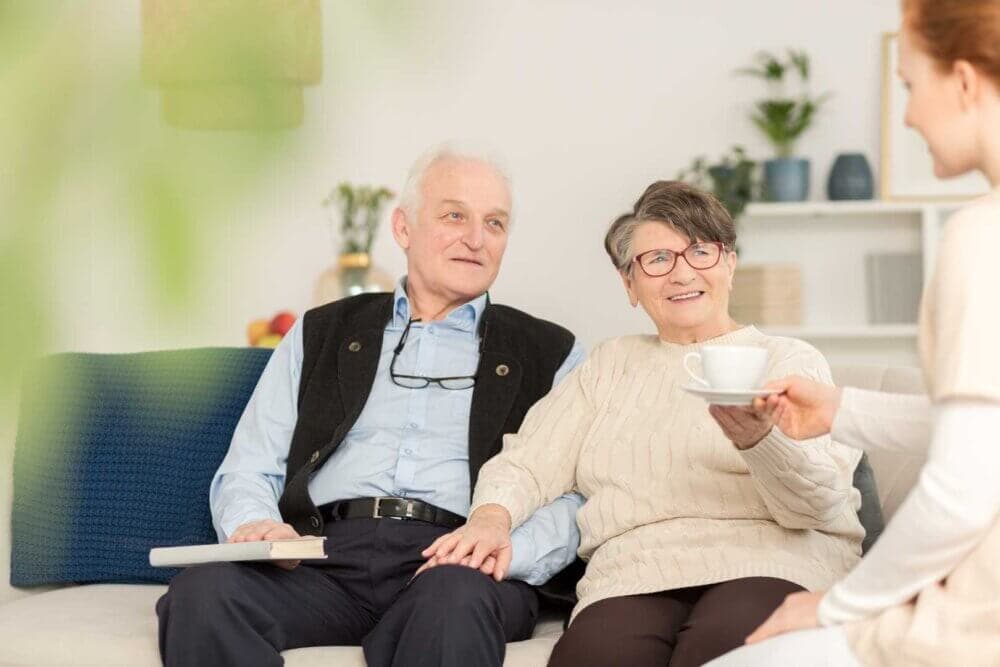 A smiling elderly couple sits on a couch, holding hands. A woman offers them a cup of tea. - Home Instead