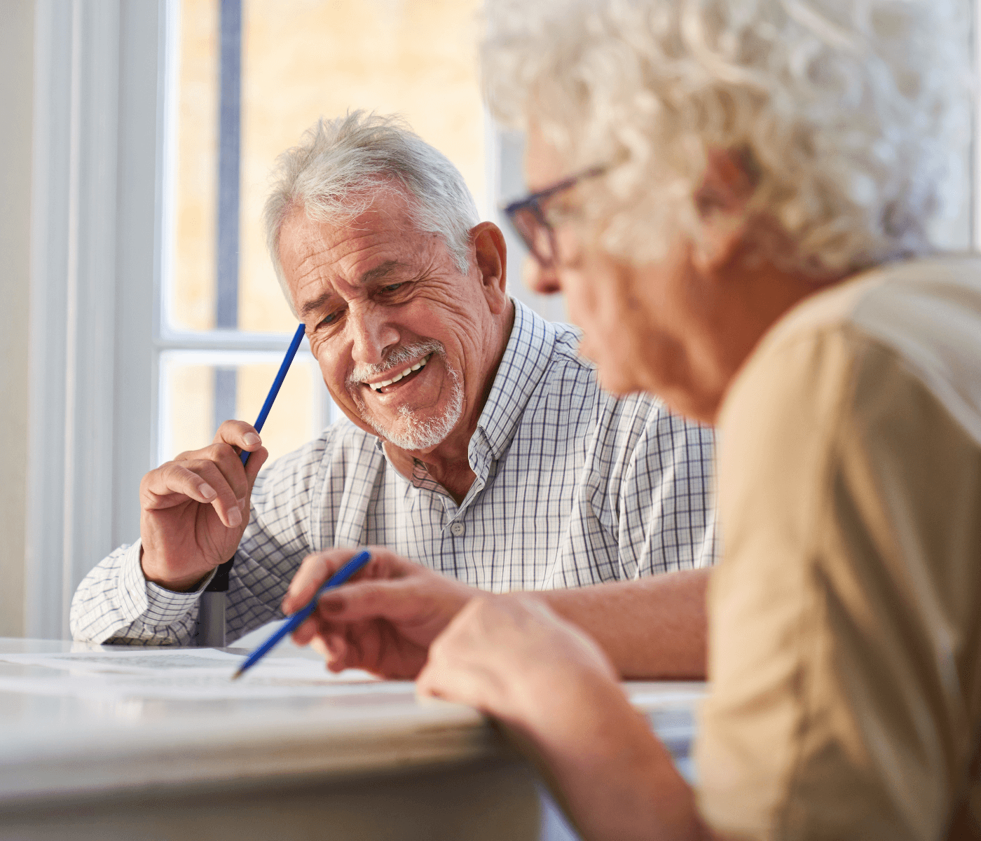 Two elderly people, one smiling and holding a pen, work on documents at a table near a bright window. - Home Instead