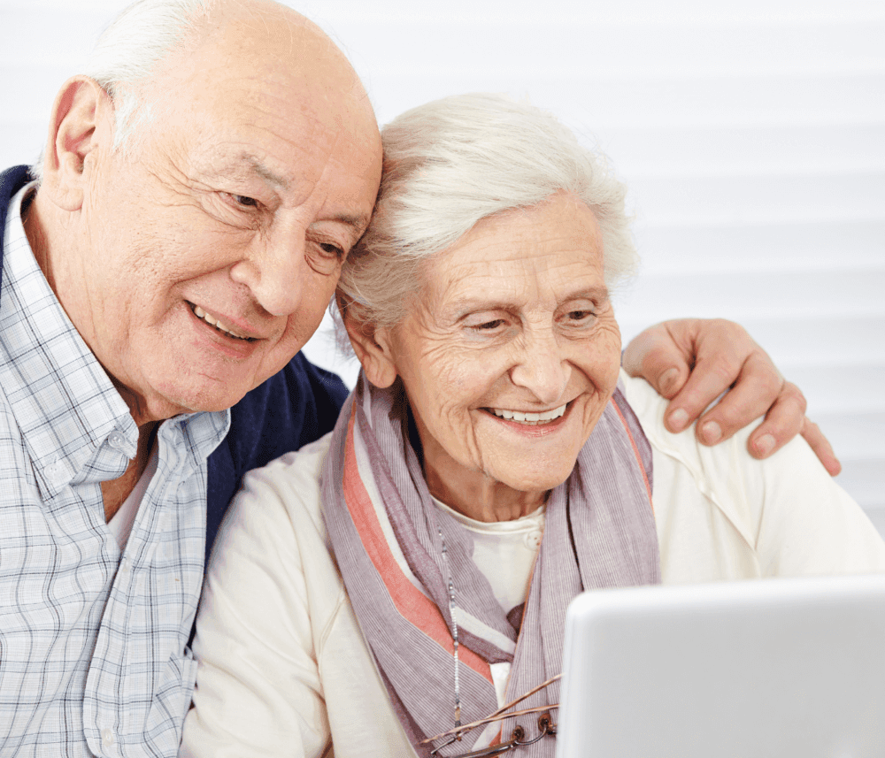 An elderly couple smiles as they look at a tablet together, sitting closely and enjoying their time. - Home Instead