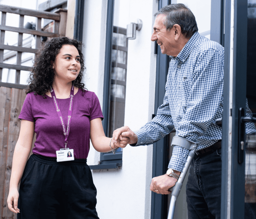 A caregiver with a name tag helps a man using crutches at the entrance of a building, holding his hand supportively. - Home Instead