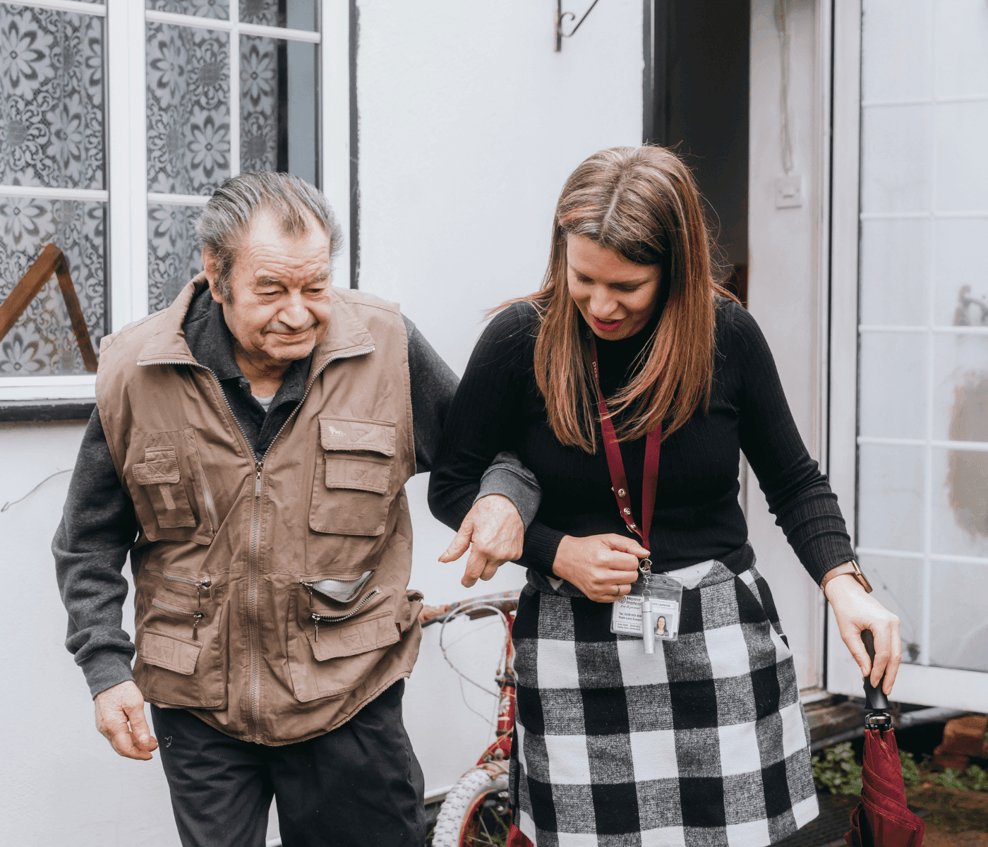 An elderly man in a brown vest walks arm-in-arm with a woman in a black top and plaid skirt outside a building. - Home Instead