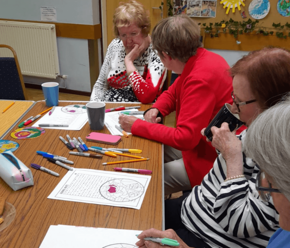 Four elderly women are seated at a table, coloring and chatting, with coloring tools and sheets spread out before them. - Home Instead