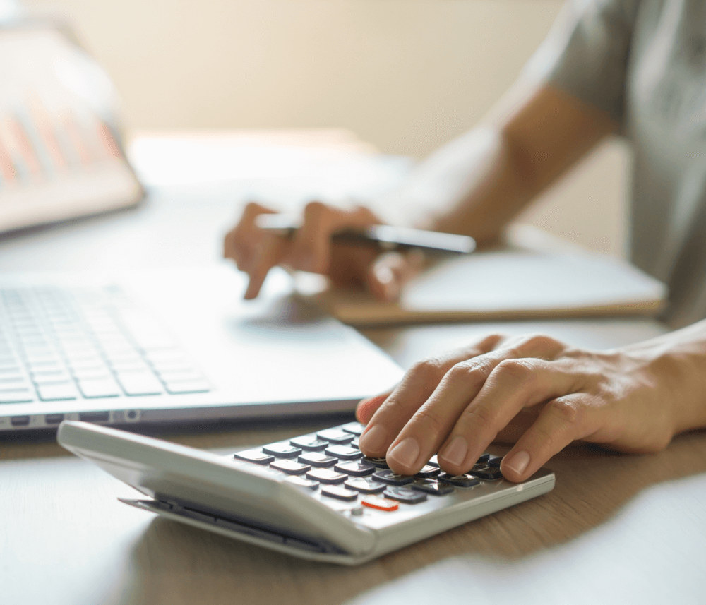 Person using a calculator and laptop while holding a pen, focusing on calculations or financial work at a desk. - Home Instead