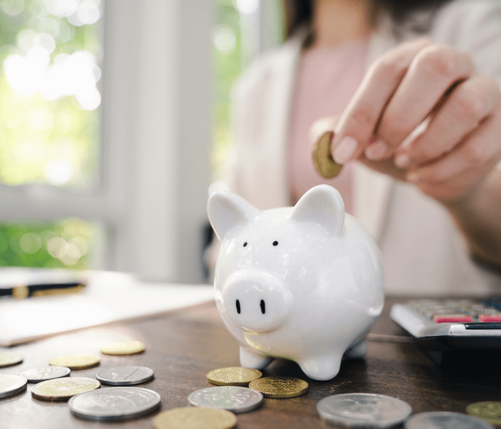 Person placing a coin into a white piggy bank on a table with scattered coins and a calculator. - Home Instead