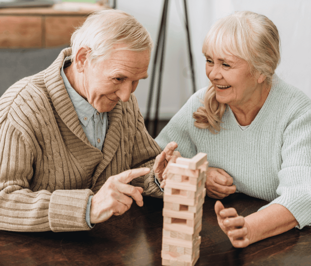Elderly couple smiling and playing a game of Jenga at a table indoors. - Home Instead