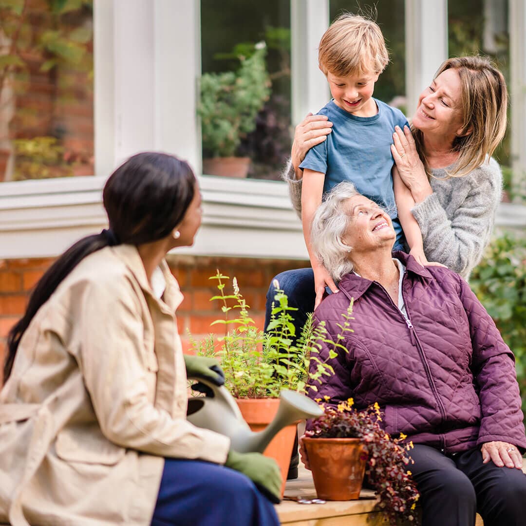 Three generations of a family enjoying time together in a garden. An elderly woman, a young boy, and two women smile. - Home Instead
