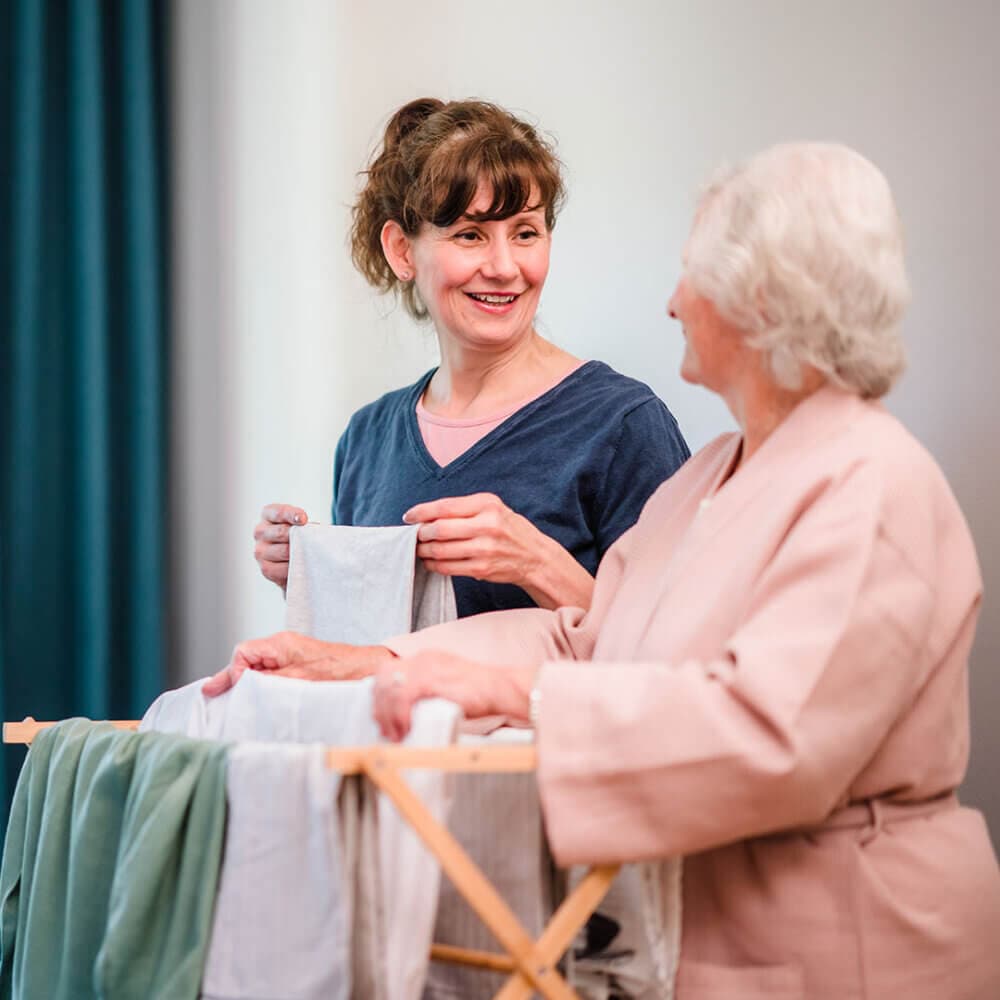 A woman smiles while helping an elderly lady hang laundry on a drying rack. - Home Instead