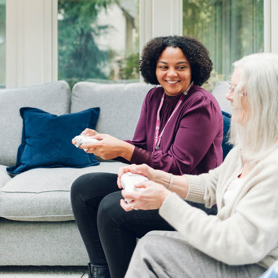 Two women sit on a sofa playing video games, one older with white hair and one younger with curly hair, both smiling. - Home Instead