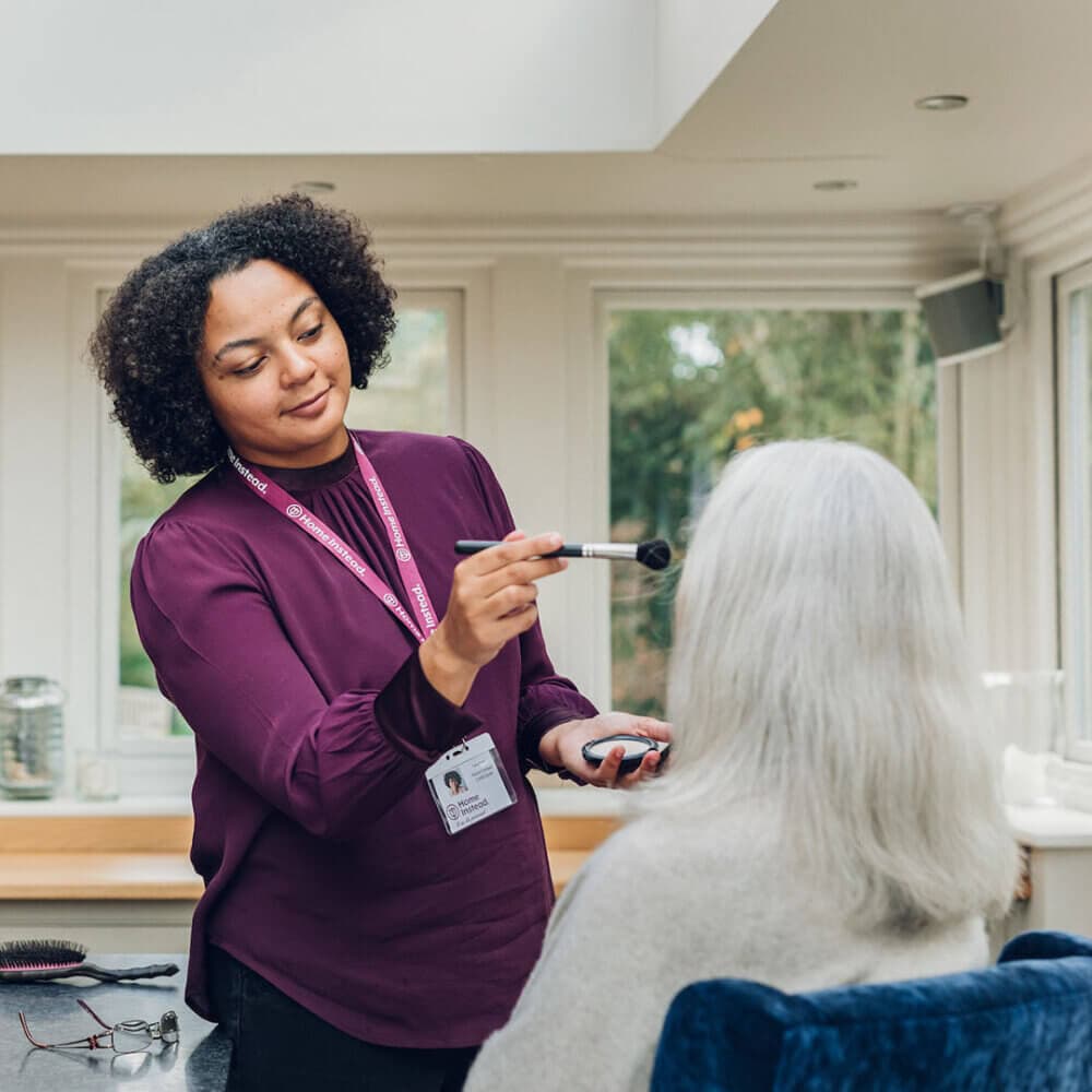 A caregiver with curly hair in a purple shirt applies makeup to an elderly person with long white hair. - Home Instead
