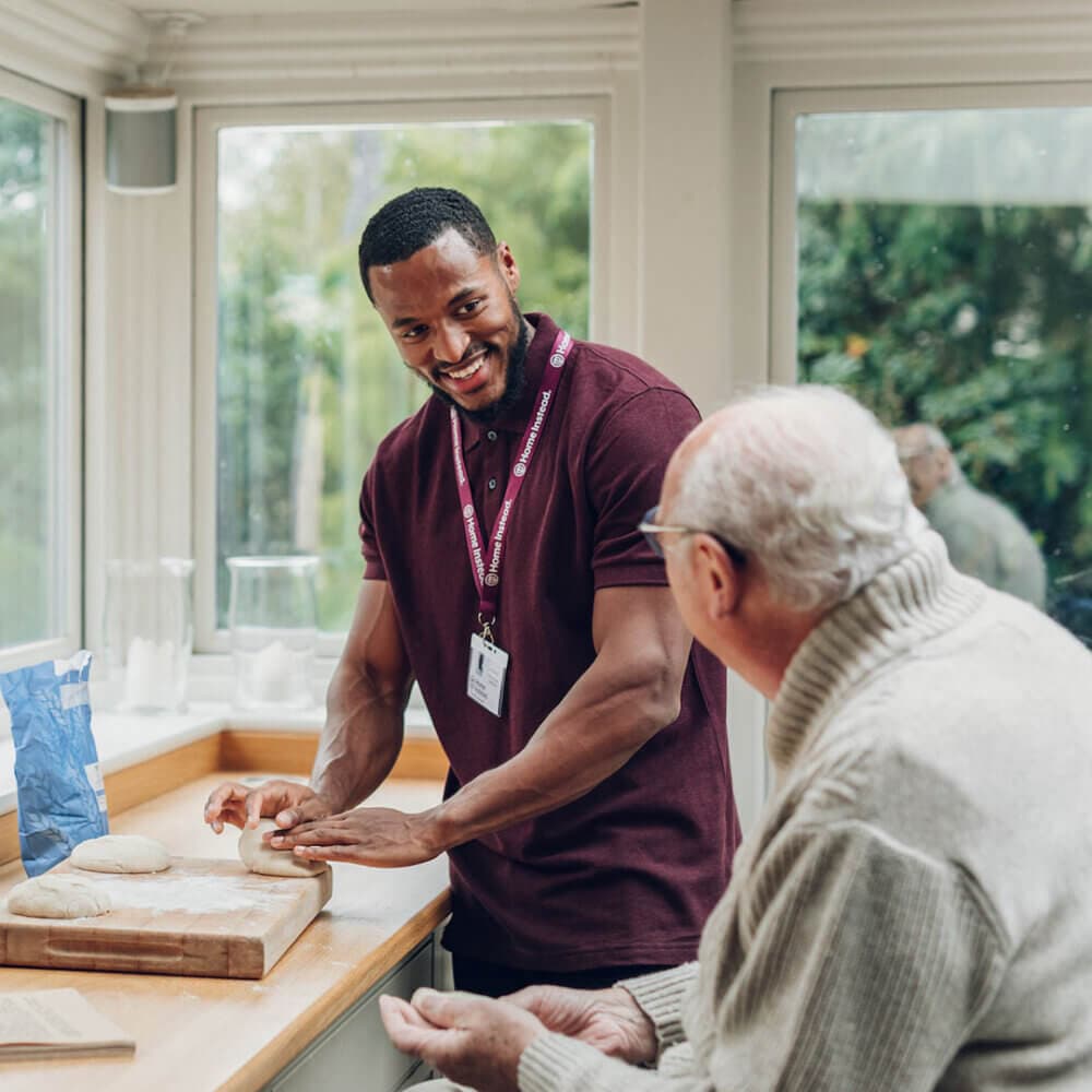 A man helps an elderly person knead dough in a kitchen with large windows and greenery outside. - Home Instead