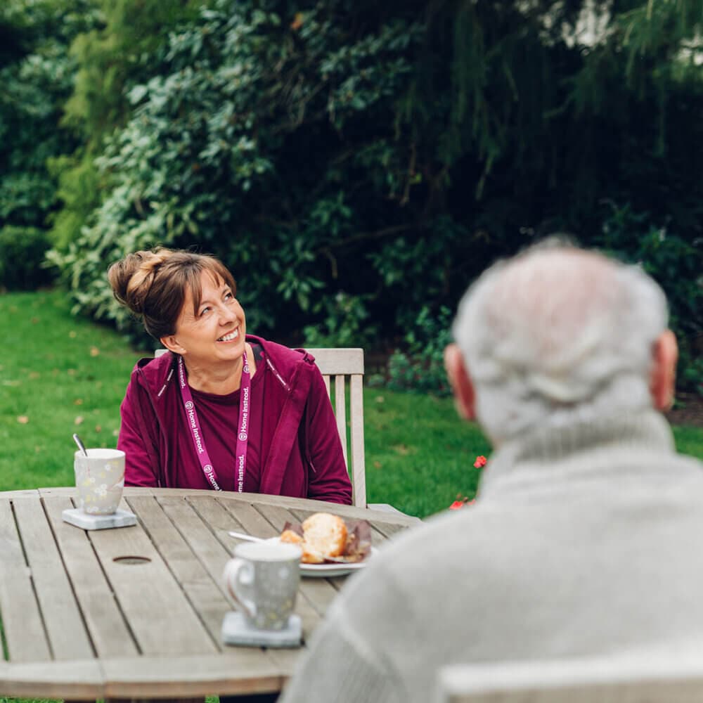 A woman smiles at an older man while sitting at a wooden table outside with cups and a dessert on the table. - Home Instead