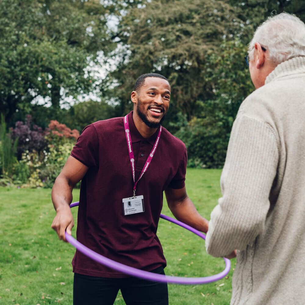 A man wearing a lanyard helps another person with a hula hoop in a garden, both smiling and enjoying the activity. - Home Instead