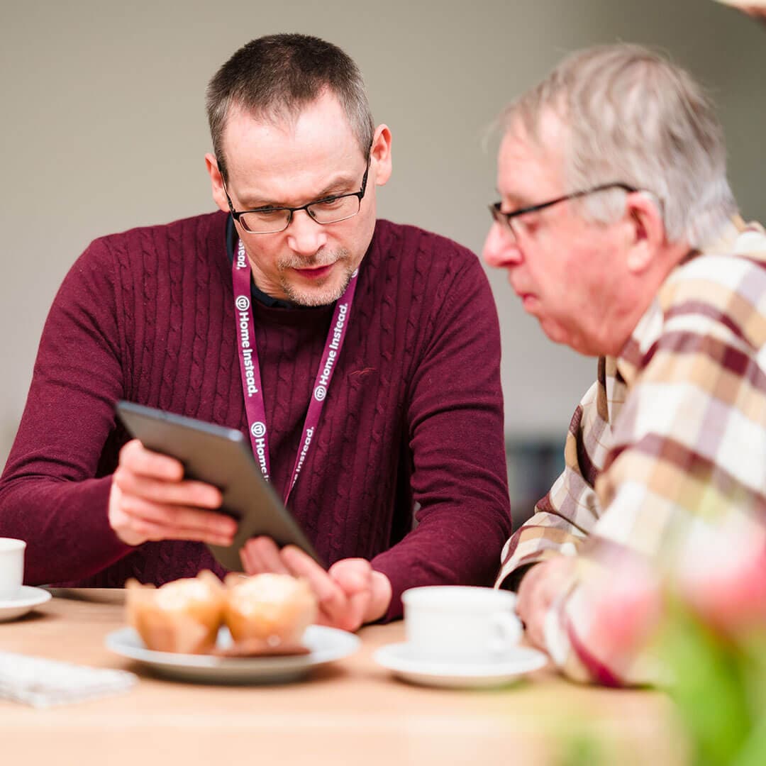 Two men sitting at a table with coffee and muffins, one showing the other something on a tablet. - Home Instead